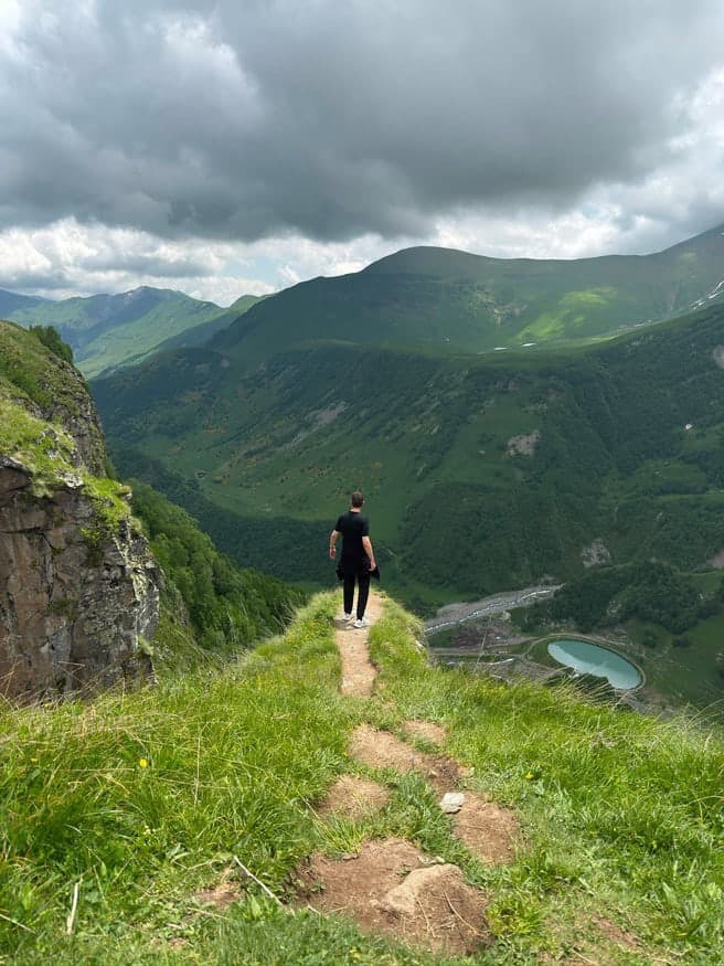 Wanderer auf einem schmalen Grasgrat, darunter ein Fluss im grünen Tal